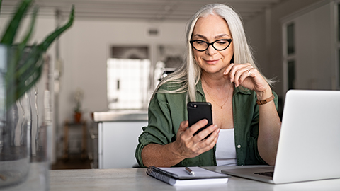 Frau sitzt zu Hause am Schreibtisch mit Laptop, Notizblock und Smartphone in der Hand, um sich über das Hilfsangebot zu informieren.